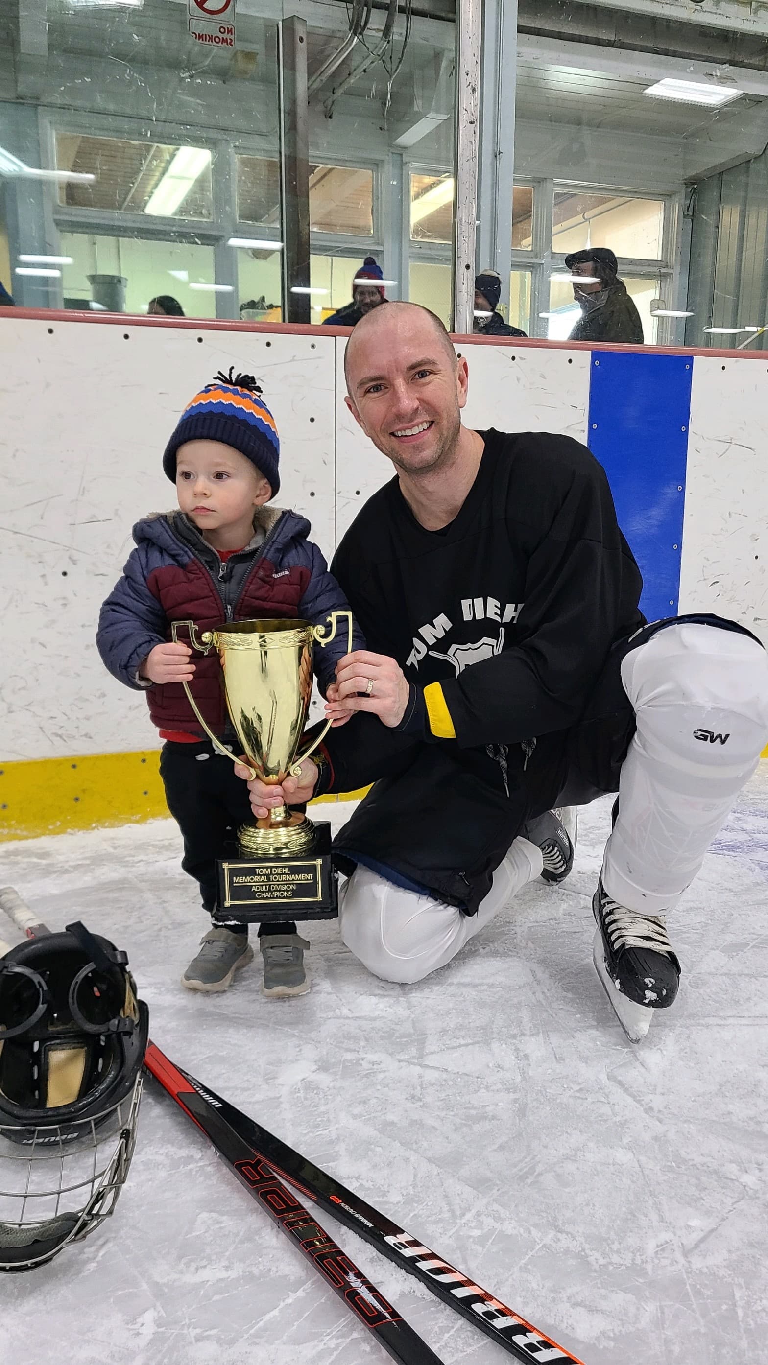 Brett in hockey gear on the ice holding a tournament trophy with a small child